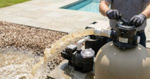 A powerful, turbulent stream of dark, dirty brown water being discharged from a pool filter system via a dedicated hose onto a gravel path.