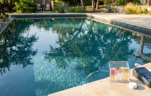 Close-up of a standard chemical test kit and digital water tester sitting on a stone pool coping. This visual connects to the section discussing how quality companies monitor chemical balance.
